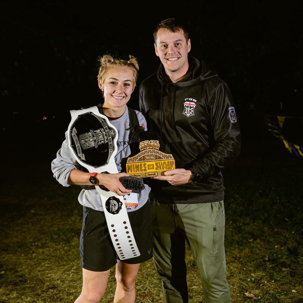 A runner and her crew member pose at night with a championship belt and first-place trophy after finishing the Mines of Spain 100-mile race.