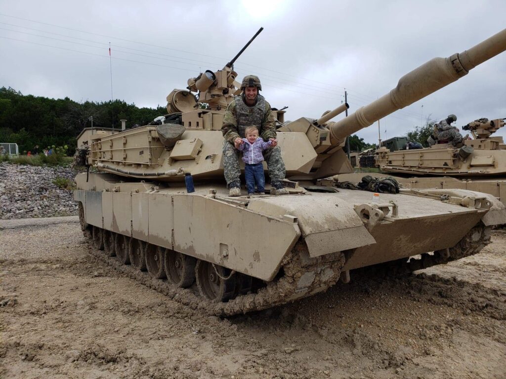 A soldier sits with a young child on top of an M1 Abrams tank at a military training area.