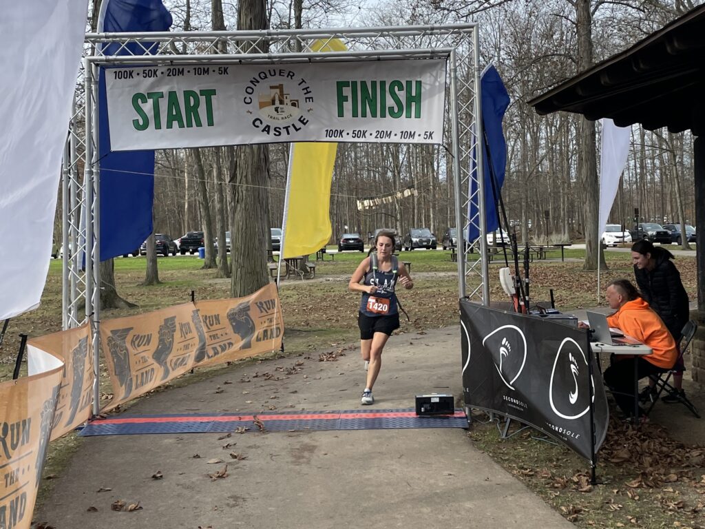 A runner crosses the finish line at the Conquer the Castle trail race beneath a start-and-finish arch.