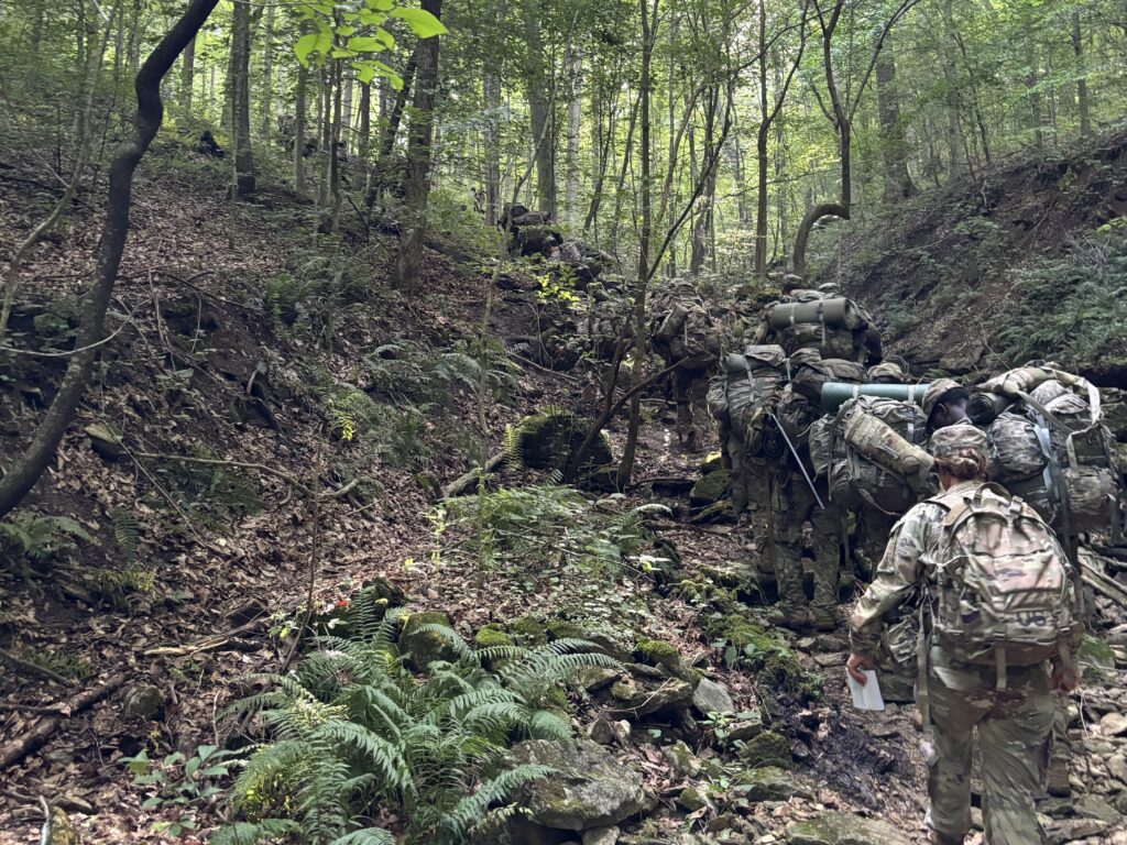Soldiers carrying heavy rucksacks move single file through a steep, wooded ravine during field training.